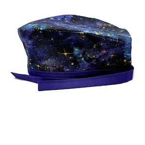 May include: A purple and blue scrub hat with a galaxy print and gold stars. The hat has a purple ribbon tie.