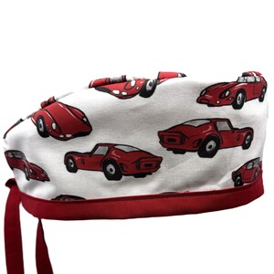 May include: White surgical scrub cap with a repeating pattern of red race cars. The cap has a red trim and ties. The cars are in various positions and angles. The background is white.