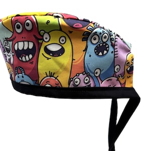 Może przedstawiać: Kolorowy czepek chirurgiczny z wzorem kreskówek potworów. Czepek ma czarną gumkę i sznurki.