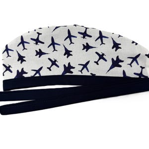 May include: A white surgical scrub cap with a navy blue airplane pattern. The cap has a navy blue band and long ties for securing. The aviation-themed cap is designed for medical professionals or anyone in the aviation field.