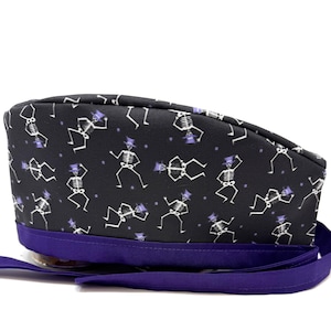 May include: A black and purple skull cap with a repeating pattern of white skeletons wearing top hats. The cap has a purple band and long purple ties. The cap is likely made of fabric.