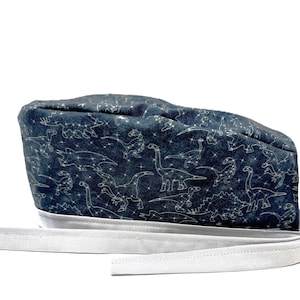 Może przedstawiać: Czapka chirurgiczna w kolorze ciemnoniebieskim z białym paskiem. Czapka ma wzór białych dinozaurów na ciemnoniebieskim tle.