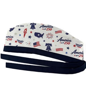 May include: A white scrub cap with a navy blue trim and ties. The cap is patterned with patriotic symbols like the Liberty Bell, flags, stars, and the text "America ***250". The design celebrates the 250th anniversary.