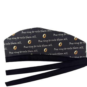 May include: A black scrub cap featuring a repeating pattern of gold-colored rings and the white text "One ring to rule them all." The cap has a solid black fabric band at the bottom and two long black fabric ties extending from the sides for fastening.