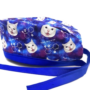 Może przedstawiać: Niebieski i fioletowy czepek chirurgiczny z powtarzającym się wzorem białych kotów i nadrukowanym słowem "Ketamina". Czepek ma niebieski sznurek.