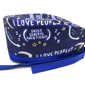 May include: Blue scrub hat with a white print that says "I love people under general anesthesia" with stars and medical symbols. The hat has a blue tie.
