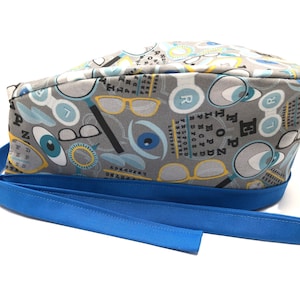 Op de afbeelding: Een grijze en blauwe chirurgische scrub-muts met een oogonderzoekkaartpatroon. De muts heeft een blauwe lintband. Het patroon bevat brillen, ogen en de letters "E", "P", "O", "R" en "D".