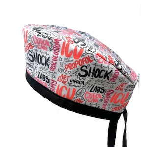 Op de afbeelding: Een witte scrub-muts met zwarte en roze medische termen erop gedrukt. De muts heeft een zwarte stropdas.