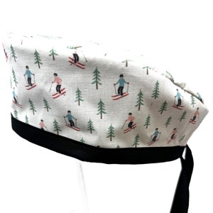 May include: White surgical scrub cap with a black band and ties. The cap features a pattern of skiers in blue and pink, and green pine trees. The cap is made of a soft, breathable fabric.