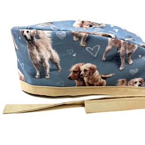 May include: A blue scrub hat with a pattern of golden retriever dogs and hearts. The hat has a tan tie.