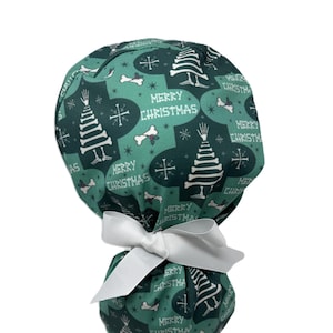 Puede incluir: Un gorro quirúrgico verde con un lazo blanco. El gorro presenta un patrón repetido de árboles de Navidad, huesos y las palabras "MERRY CHRISTMAS" en blanco. El gorro está atado en la parte posterior con un lazo blanco.