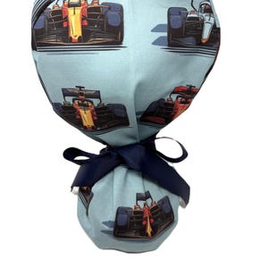 May include: A light blue scrub cap featuring a repeating pattern of orange and black Formula 1 race cars. The cap is secured at the back with a dark navy blue grosgrain ribbon tie, creating a gathered effect at the base of the head covering.