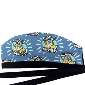 May include: A blue scrub cap featuring a repeating graphic print of a hand slapping a yellow object with the text "SLAP IT HARDER!" in white and black lettering. The cap has a solid black fabric band at the bottom with two long black tie straps.