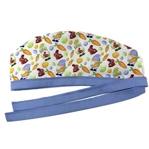 May include: A white Easter-themed scrub cap with a repeating pattern of gnomes, chocolate bunnies, carrots, and Easter eggs. The cap has a light blue trim and long ties for securing. This cap is designed for medical professionals.