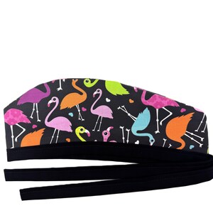Peut inclure: Une charlotte chirurgicale noire ornée d'un motif coloré de flamants roses dans des tons de rose, orange, vert lime et bleu clair, agrémenté de petits cœurs. Elle possède une bande en tissu noir à la base et de longs liens noirs à nouer.