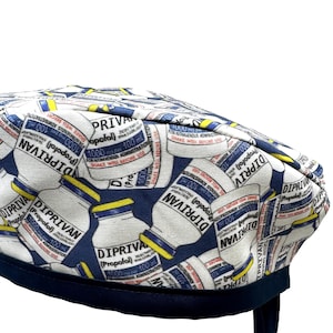 May include: A white and navy blue scrub cap featuring a repeating pattern of medicine bottles with the text "DIPRIVAN (Propofol)". The cap has a navy blue band and ties.