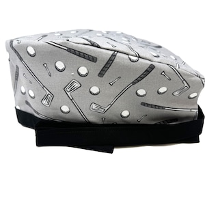 Op de afbeelding: Grijze scrub-muts met een zwart lint. De muts heeft een herhalend patroon van golfclubs, golfballen en tees.