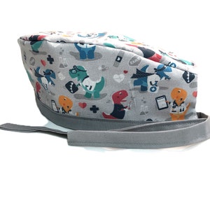 Op de afbeelding: Grijze scrub-muts met een kleurrijk dinosaurusprint met dinosaurussen die dokterskostuums en medische symbolen dragen. De muts heeft een grijze stropdas.