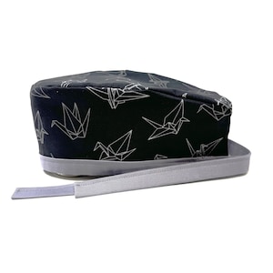 Op de afbeelding: Een zwarte scrub-muts met een wit origami-kraanvogelpatroon en een lichtgrijze verstelbare strop.