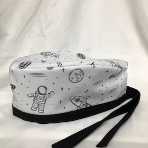 Może przedstawiać: Biały chirurgiczny czepek scrub z czarną lamówką i nadrukiem w kosmicznym stylu przedstawiającym astronautów, planety, gwiazdy i rakiety. Czepek ma czarne sznurki.