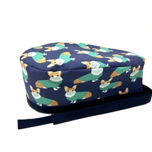 May include: A blue scrub hat with a pattern of corgis wearing surgical masks and scrubs. The hat has a navy blue adjustable tie.