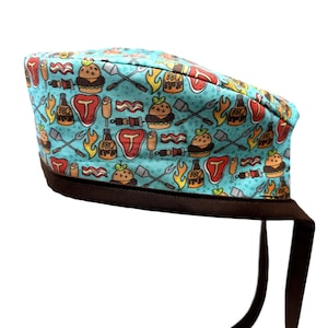 May include: A turquoise blue scrub hat with a brown elastic band and tie. The hat has a repeating pattern of cartoon images of grilling food, including burgers, hot dogs, steak, corn on the cob, and barbecue sauce.