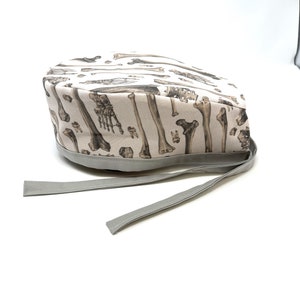 Op de afbeelding: Een witte scrubmuts met een bottenprint en een grijze stropdas. De muts is gemaakt van een zachte, comfortabele stof en is perfect voor medische professionals.