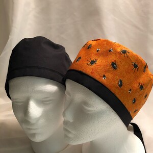 May include: Two scrub caps on white mannequin heads. One is solid black, the other is orange with a black spider and web pattern. Both have a black band around the base and ties in the back.