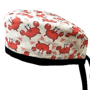 May include: A patterned scrub cap featuring a repeating design of red cartoon crabs on a beige background. The cap has a black band and ties. The crabs have large eyes and are set against a background of small, stylized seaweed.