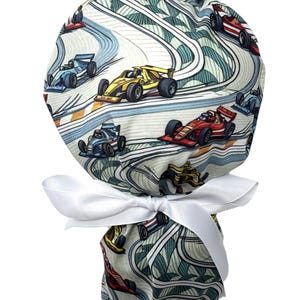 May include: A scrub cap featuring a repeating pattern of colorful race cars on a track with blue, yellow, and red vehicles. The fabric has a light background with wavy lines and is gathered at the back with a plain white ribbon tied in a bow.