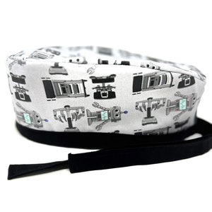 Op de afbeelding: Een witte scrub-muts met een zwarte stropdas. De muts heeft een herhalend patroon van robots met moersleutels en printplaten.