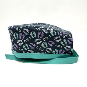 May include: A black scrub hat with a teal ribbon. The hat is covered in a pattern of purple, green, and white footprints with the text "L&D" repeated throughout.