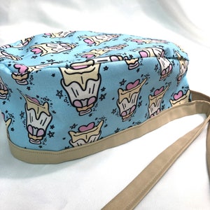 Może przedstawiać: Jasno niebieski czepek chirurgiczny z brązowym paskiem. Czepek ma powtarzający się wzór kreskówek z babeczkami z różowymi sercami i gwiazdami.