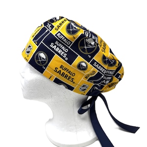 Puede incluir: Un gorro quirúrgico amarillo, azul marino y blanco de los Sabres de Buffalo. El gorro presenta el logotipo del equipo, su nombre y el logotipo de la NHL. El gorro tiene lazos azul marino para asegurarlo. El gorro se muestra en una cabeza de maniquí blanca.