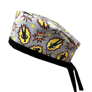 Op de afbeelding: Een grijze en zwarte scrub-muts met een superhelden-thema. De muts heeft een zwarte band en strikken. De stof heeft een herhalend patroon van gele en rode comic-stijl explosies met de woorden "HERO", "ZAP!" en "WHOOSH!" erop gedrukt. Zwarte silhouetten van superhelden bevinden zich in het midden van elke explosie.