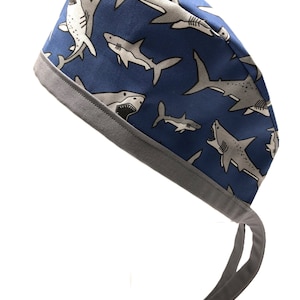 Op de afbeelding: Een blauwe chirurgische scrub-muts met een grijze rand en banden. De muts heeft een herhalend patroon van witte haaien op een blauwe achtergrond.