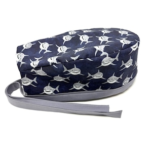May include: A blue and white surgical scrub hat with a pattern of white sharks on a dark blue background. The hat has a gray tie.
