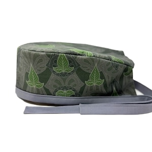 Op de afbeelding: Een groene en grijze gepatroneerde chirurgische scrub-muts met een grijze stropdas. Het patroon bevat gestileerde bladeren.