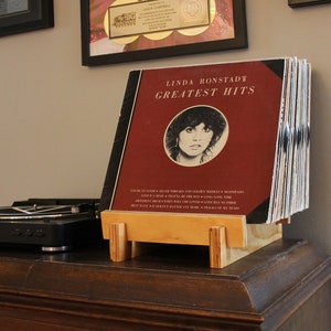 Può includere: Una pila di dischi in vinile su un supporto in legno. Il disco in cima è un album di Linda Ronstadt Greatest Hits con una copertina rossa e una foto della cantante.