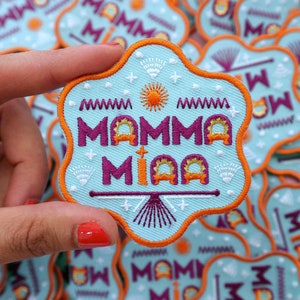 May include: Embroidered patch with the words "MAMMA MIA" in purple, outlined in orange, on a light blue background. The patch has an orange border and decorative elements including a sun, stars, and geometric patterns. The patch is held by a hand.