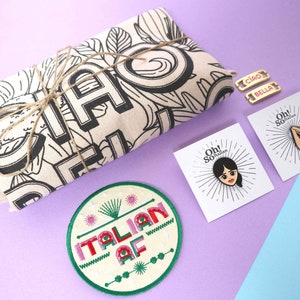 Pode incluir: Um pacote de presente embrulhado em barbante marrom e uma bolsa de lona branca com ilustrações em preto e branco. A bolsa de lona tem a palavra "Ciao" impressa. O pacote também inclui um remendo bordado verde e branco que diz "Italian AF", duas etiquetas de metal dourado que dizem "Ciao" e "Bella", e dois pinos de esmalte com o texto "Oh! So Italian" e uma imagem de desenho animado de uma mulher e um gesto de mão.