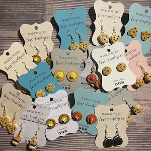May include: A collection of handmade polymer clay earrings featuring various baked goods, such as cookies, cupcakes, and s'mores. Each pair of earrings is displayed on a card with the text "Baked with love + kindness" and the artist's signature.