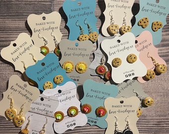 May include: A collection of handmade polymer clay earrings featuring various baked goods, such as cookies, cupcakes, and s'mores. Each pair of earrings is displayed on a card with the text "Baked with love + kindness" and the artist's signature.