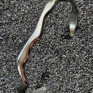 May include: A silver-colored, abstract bracelet with a curved, organic design. The bracelet has a smooth, polished finish and a unique shape. The bracelet is displayed on a dark, textured surface.