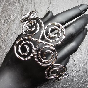 Może przedstawiać: Srebrzysta metalowa bransoletka typu mankiet z powtarzającym się wzorem połączonych ze sobą okrągłych spirali. Powierzchnia metalu ma na całym wzorze teksturowane, karbowane wykończenie, co nadaje jej metaliczny i odblaskowy wygląd.