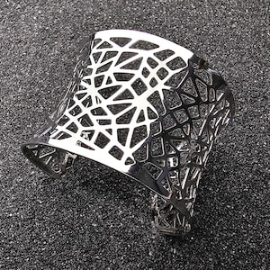 Op de afbeelding: Een zilverkleurige manchetarmband met een geometrisch openwerkontwerp. De armband heeft een brede, gebogen vorm en een gepolijste afwerking. Het openwerkpatroon bestaat uit onderling verbonden vormen, wat een modern accessoire creëert.