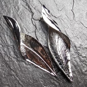 Peut inclure: Une paire de boucles d'oreilles en métal argenté en forme de feuilles allongées et torsadées. La surface de chaque boucle d'oreille présente une finition polie et réfléchissante avec des motifs de nervures gravés le long du centre et des côtés.