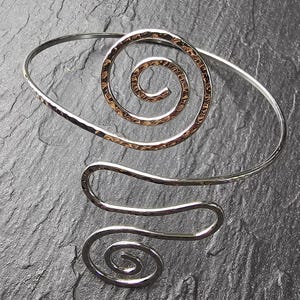 Könnte beinhalten: Silberfarbener Armreif mit einem gehämmerten, goldfarbenen Spiraldesign. Der Armreif hat ein fließendes, offenes Design mit einem zweiten, kleineren Spiraldetail. Der Schmuck wird auf einer dunklen, strukturierten Oberfläche präsentiert.