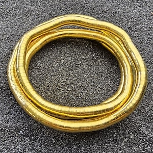 Könnte beinhalten: Ein goldfarbenes Schlangenkettarmband mit einem flexiblen, segmentierten Design, das eine durchgehende kreisförmige Schlaufe bildet. Die metallische Oberfläche hat ein poliertes Finish, das Licht reflektiert und ein glattes, gleichmäßiges Erscheinungsbild erzeugt.