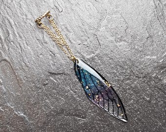 May include: A gold-toned necklace with a pendant shaped like a wing. The wing pendant features a black outline with a gradient of blue, purple, and gold glitter. The necklace is displayed on a dark gray, textured surface.